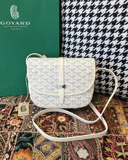A white monogrammed crossbody bag with a long strap, standing on a patterned rug