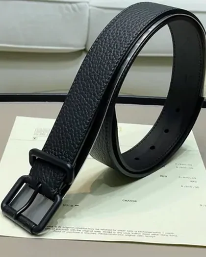 A black grained leather belt coiled upright on a white surface, buckle facing the camera